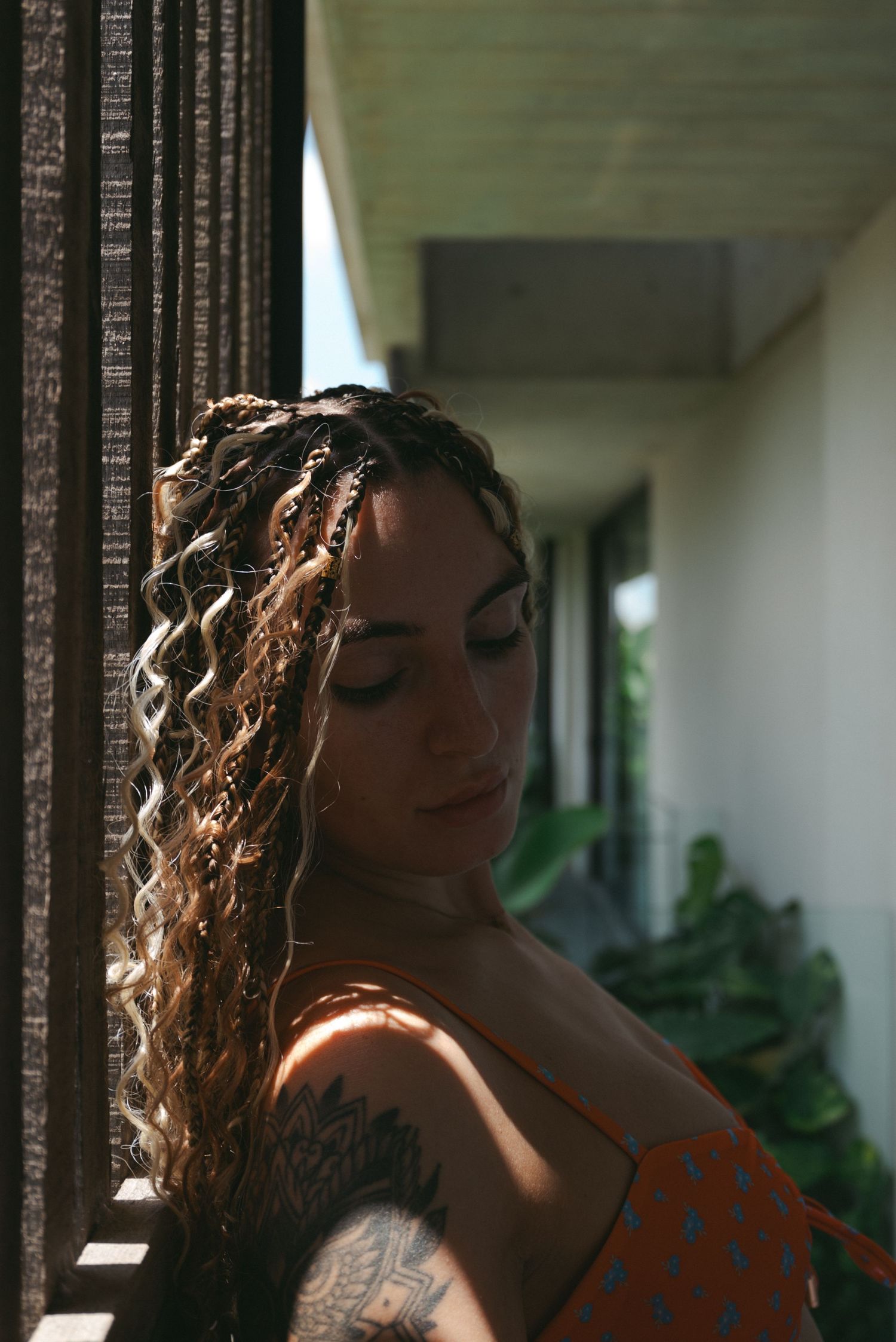 A person with curly hair and tattoos poses near a window in natural lighting wearing an orange top.