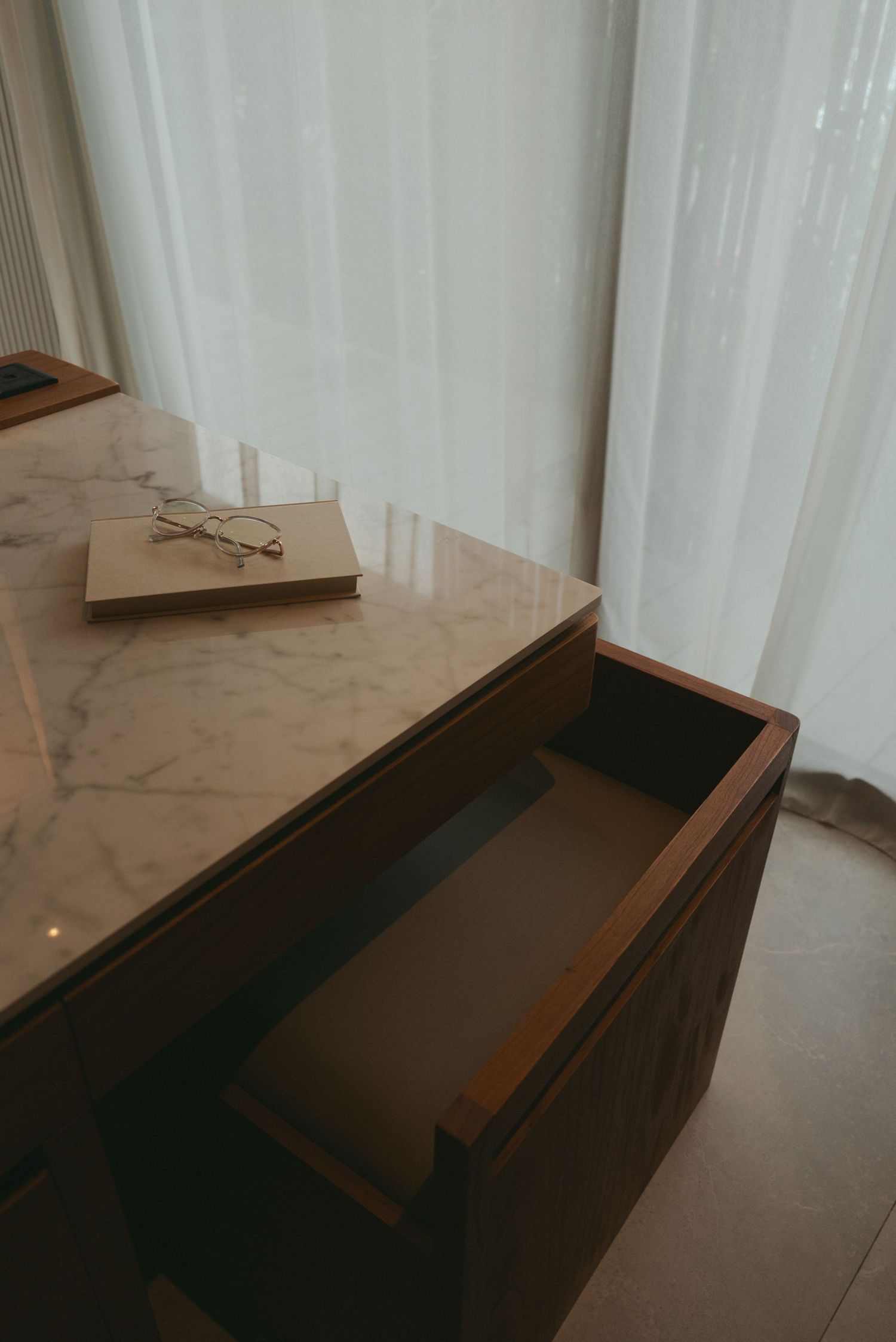 Modern wooden dresser with glossy marble top surface against white curtained wall.