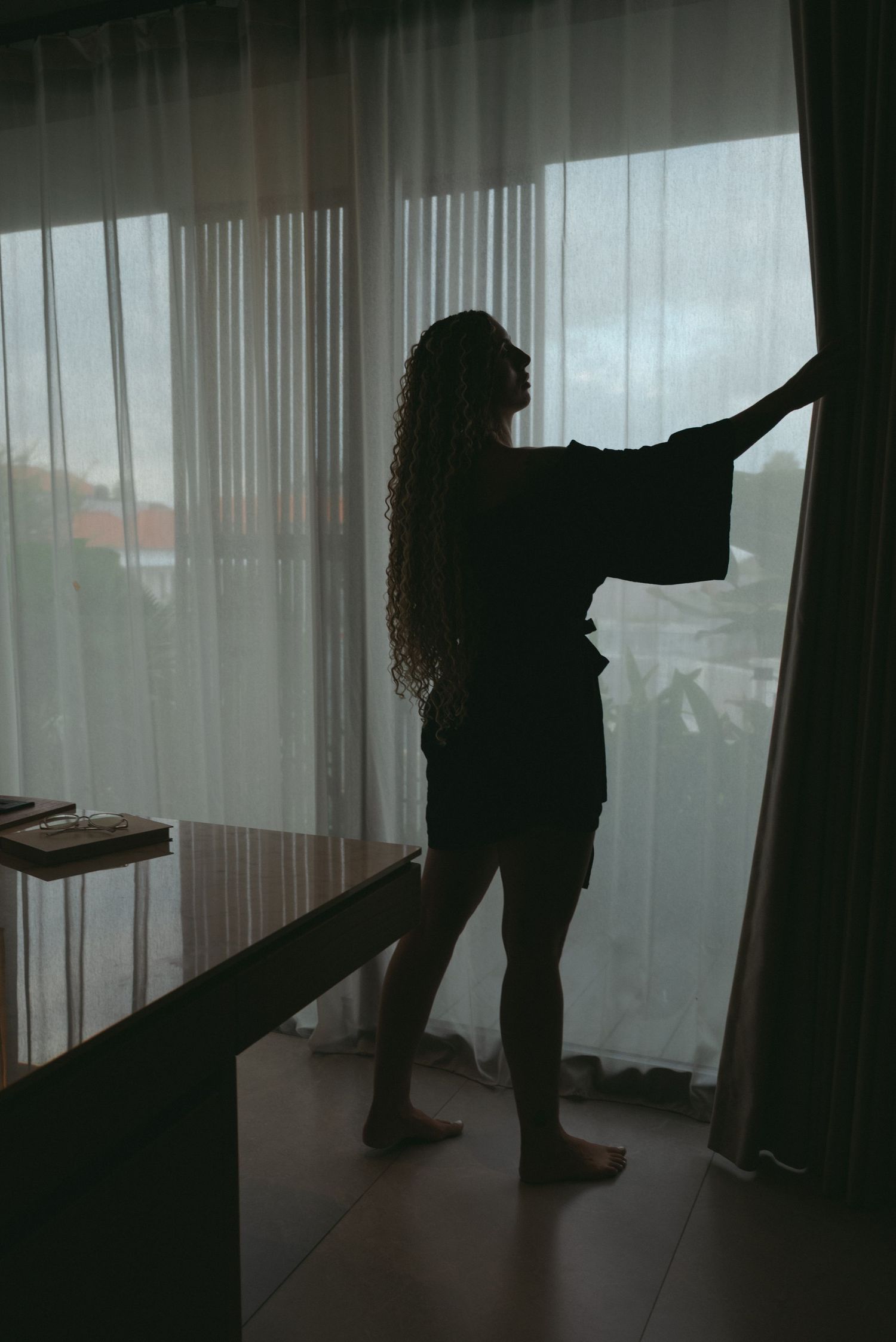 Silhouettes of figures standing behind sheer curtains in a dimly lit room create an atmospheric mood.