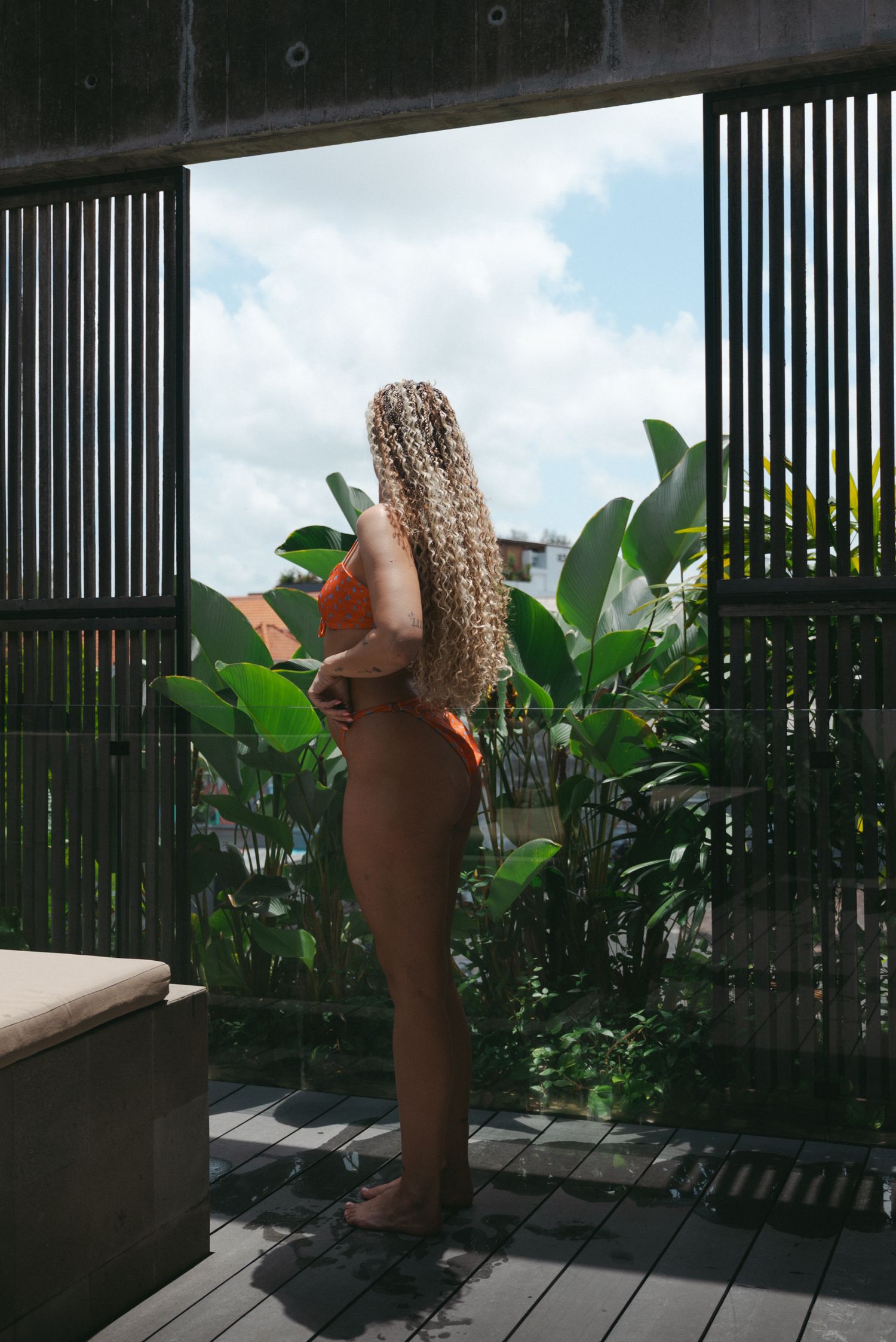 Legs in sandals and straw hat relaxing on a modern balcony with vertical slats and tropical plants.