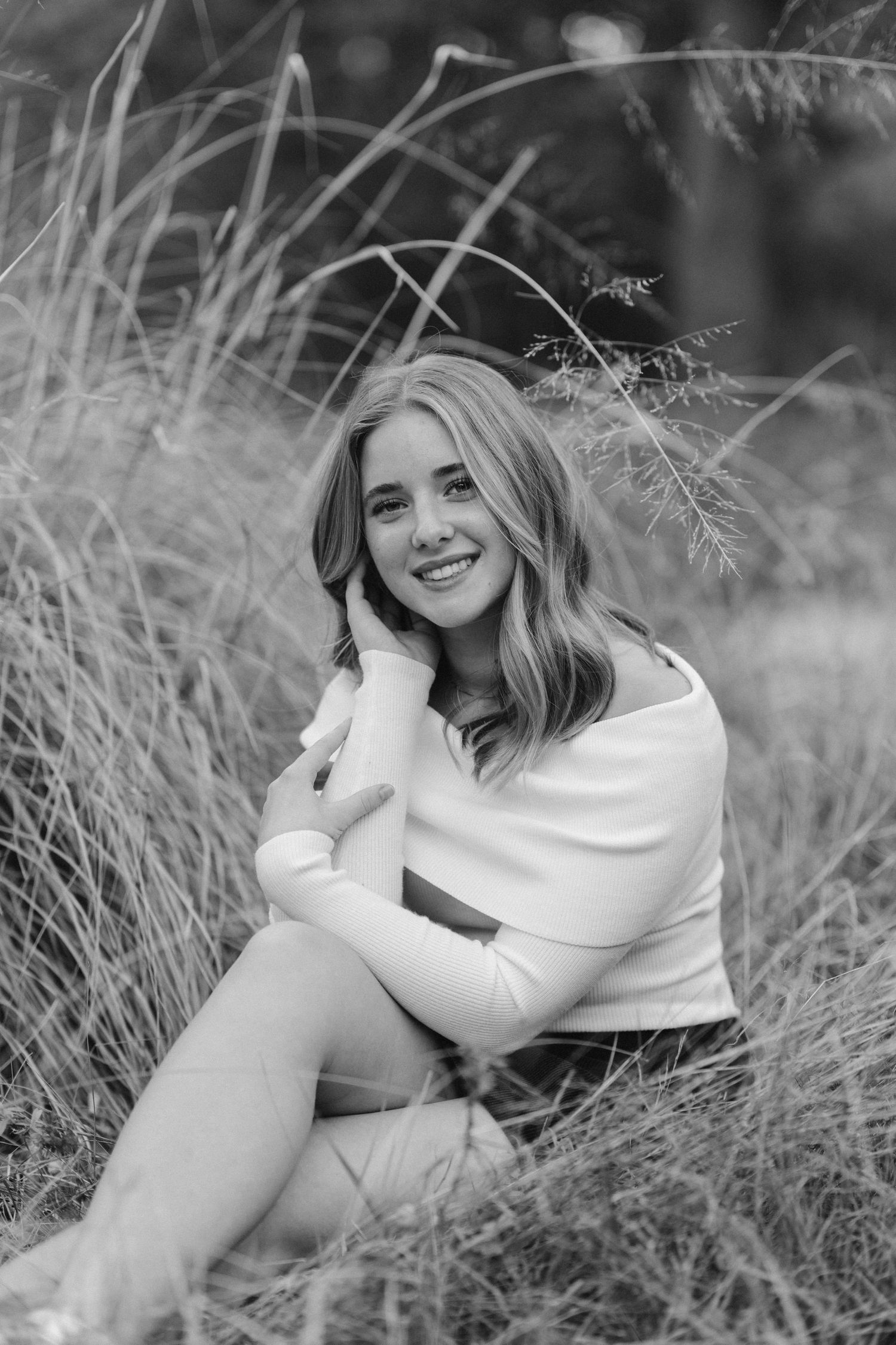 Black and white portrait of someone sitting in tall grass wearing a white sweater, smiling naturally in an outdoor setting.