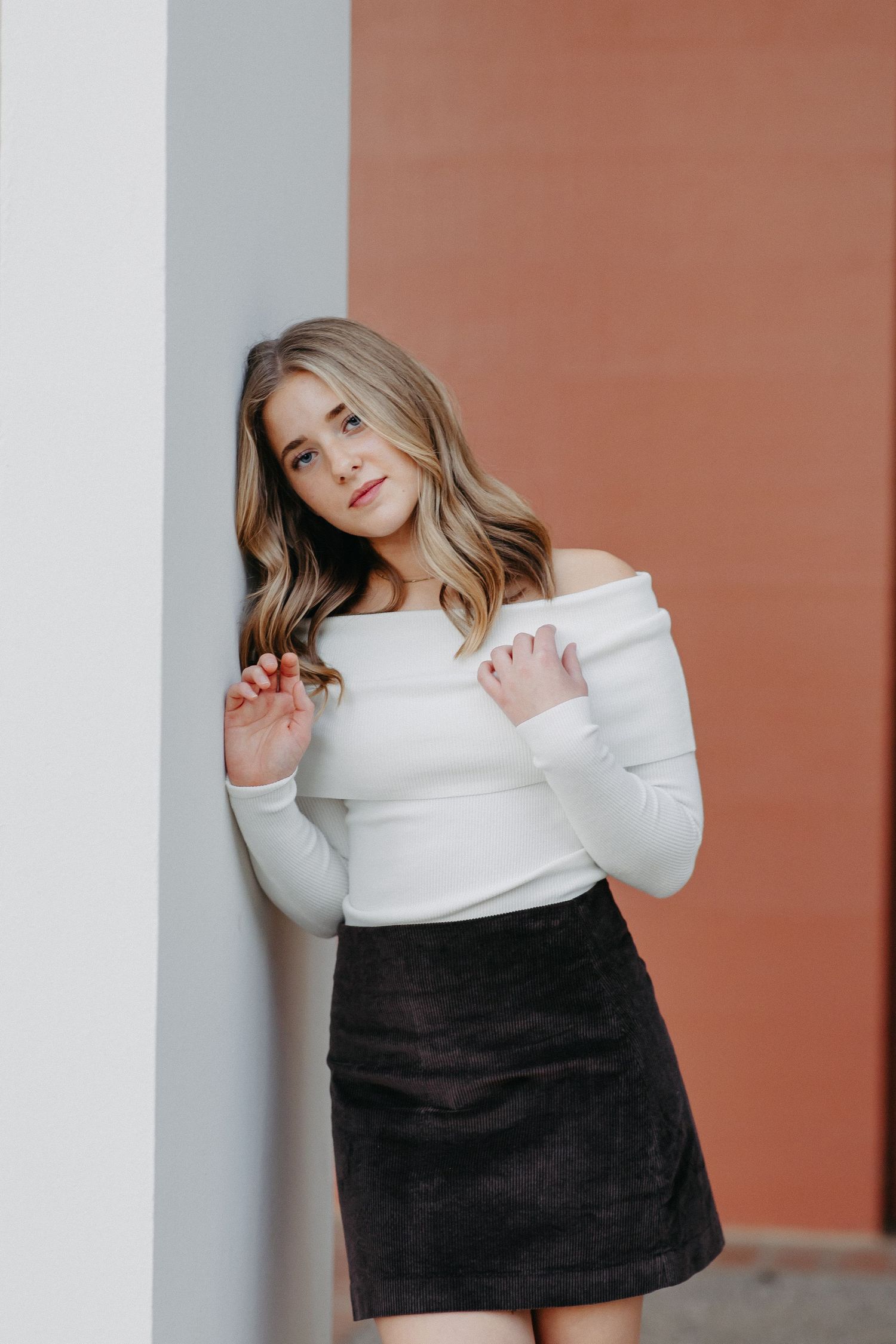 A person poses in a white off-shoulder sweater and black skirt against a coral-colored wall.