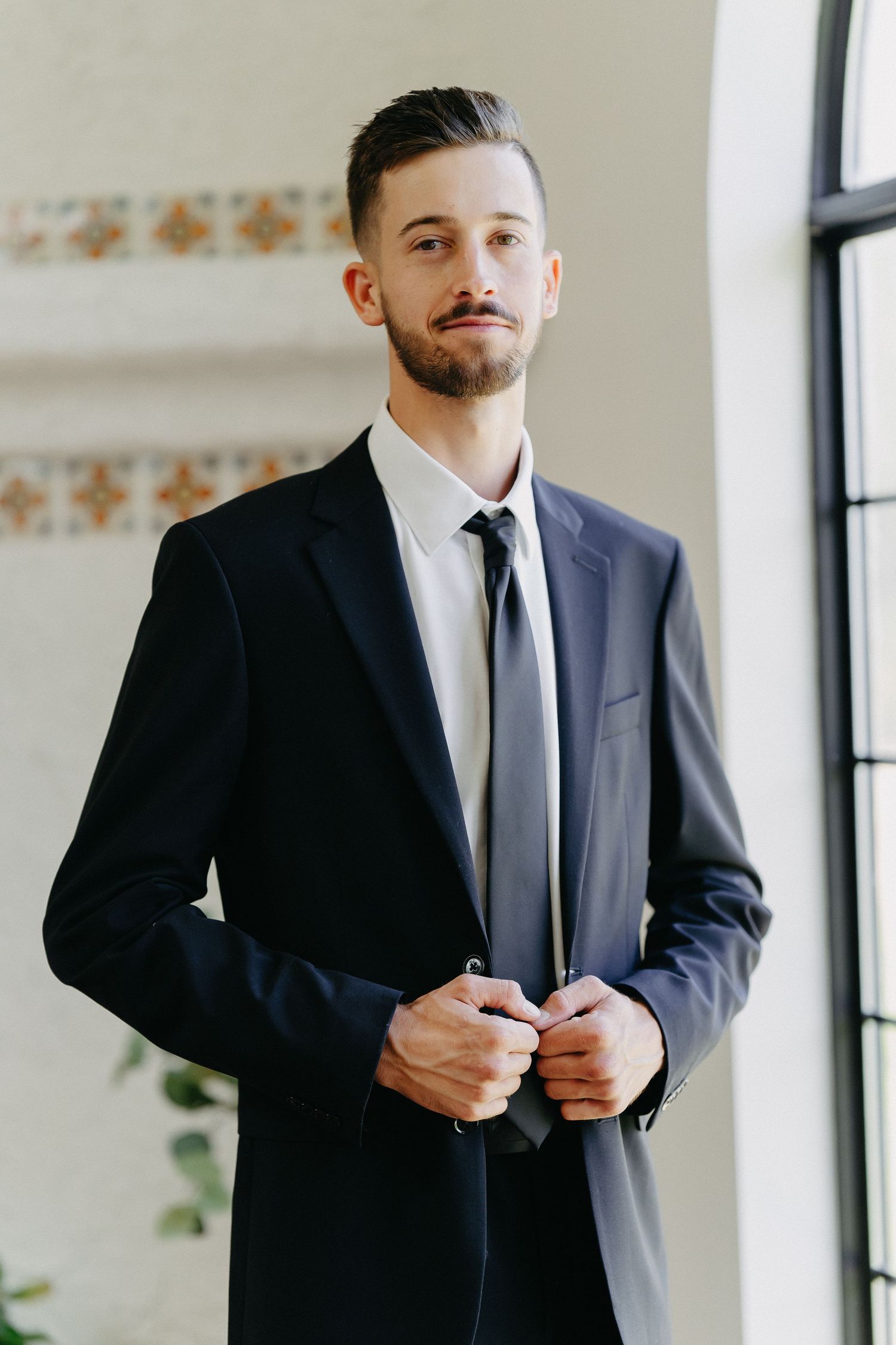 A well-dressed person in a dark suit and tie stands confidently by a window in a sunlit room.