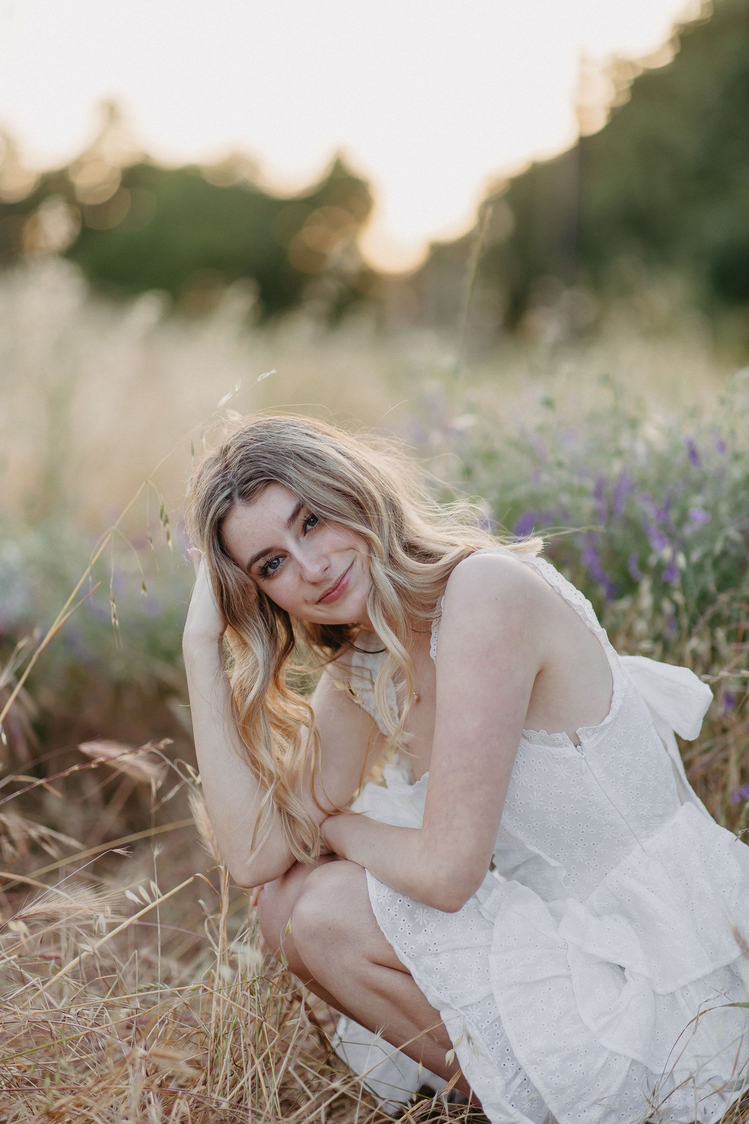 Ethereal portrait of a person in a white tiered dress sitting among wildflowers at golden hour in a meadow.