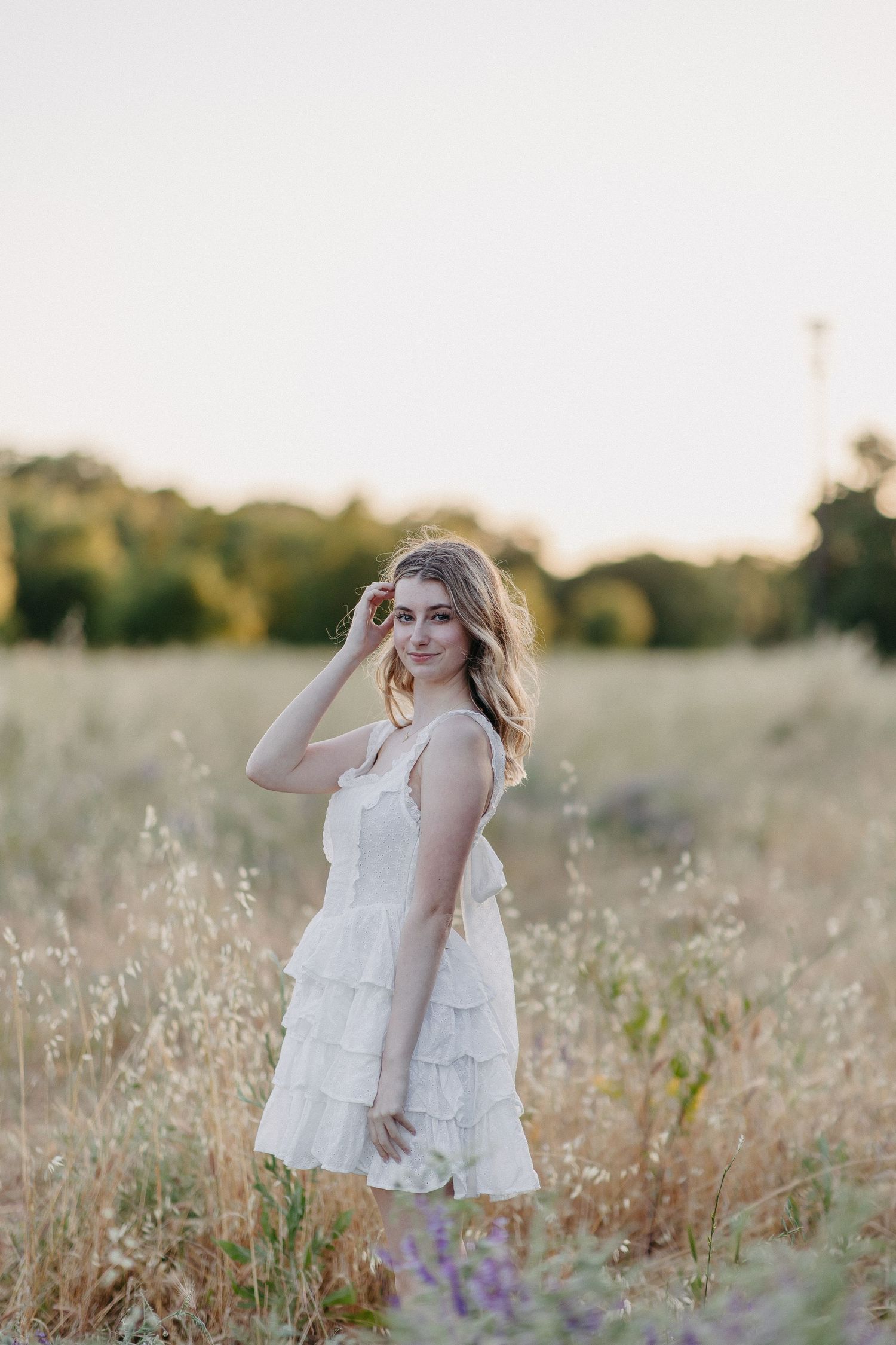 Dreamy outdoor portrait of a person in a flowing white summer dress standing in a sunlit field with purple wildflowers.