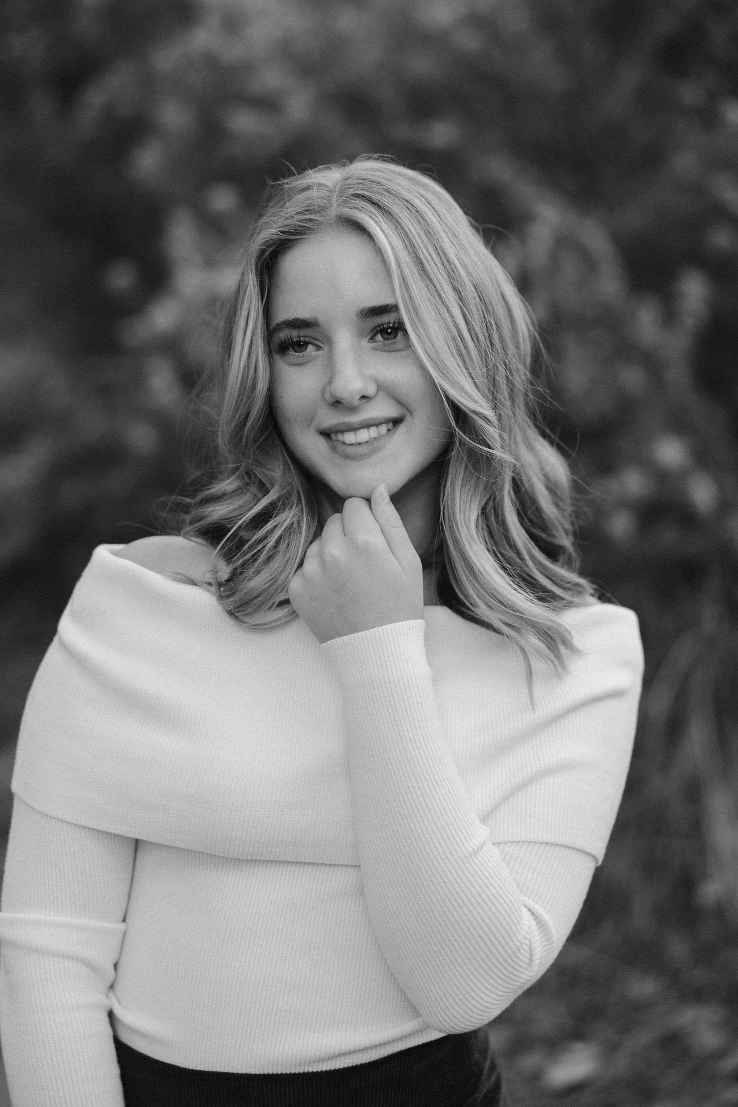A black and white portrait of a smiling person wearing a white sweater against a blurred natural background.
