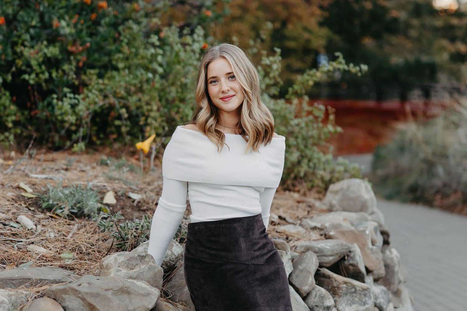 Professional outdoor portrait of someone in a white sweater and black skirt sitting on rocks in a garden setting.