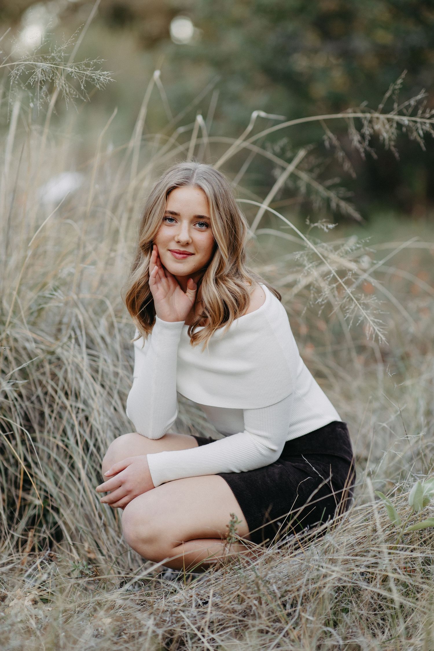 Portrait in tall grass showing woman in white sweater and black shorts crouching in autumn field.