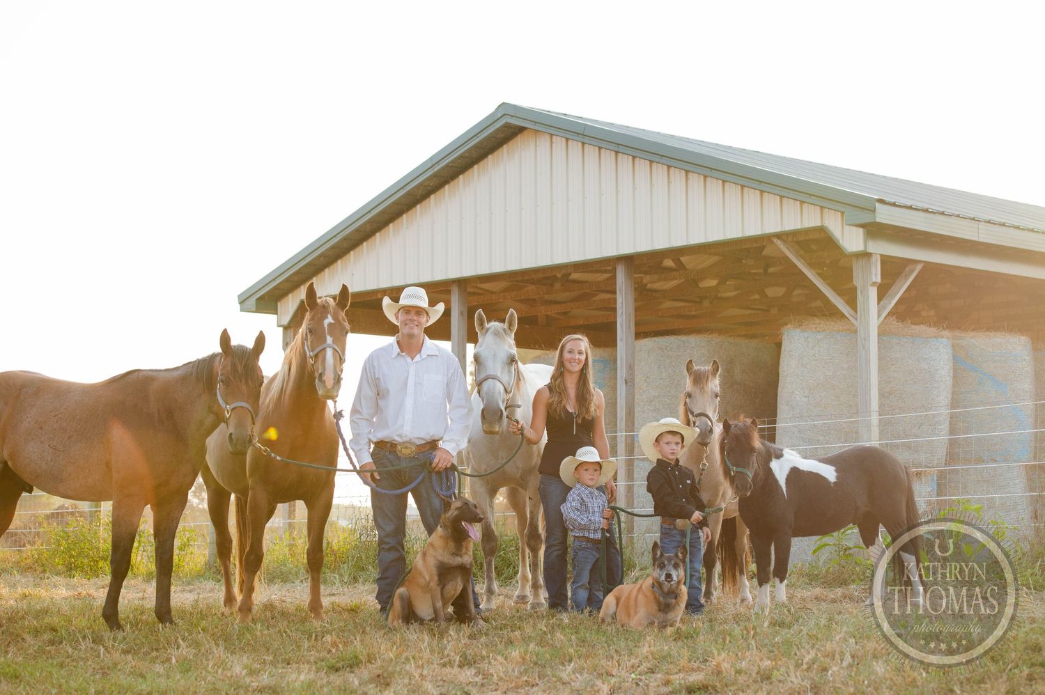 Family and pets pose together with horses outside a wooden barn at sunset.