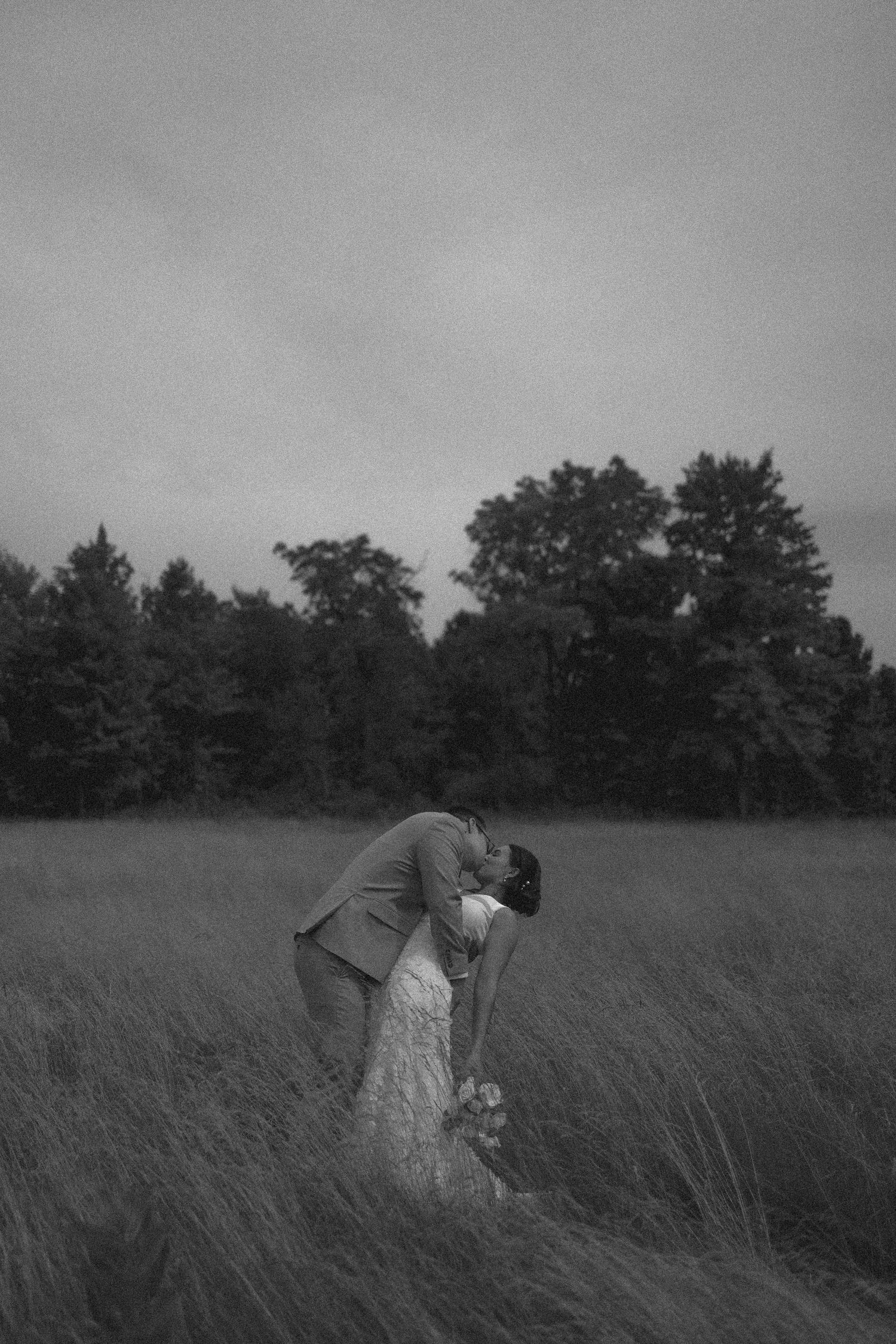 A romantic black and white photograph of a couple embracing in a misty field with tall grass and trees in the background.