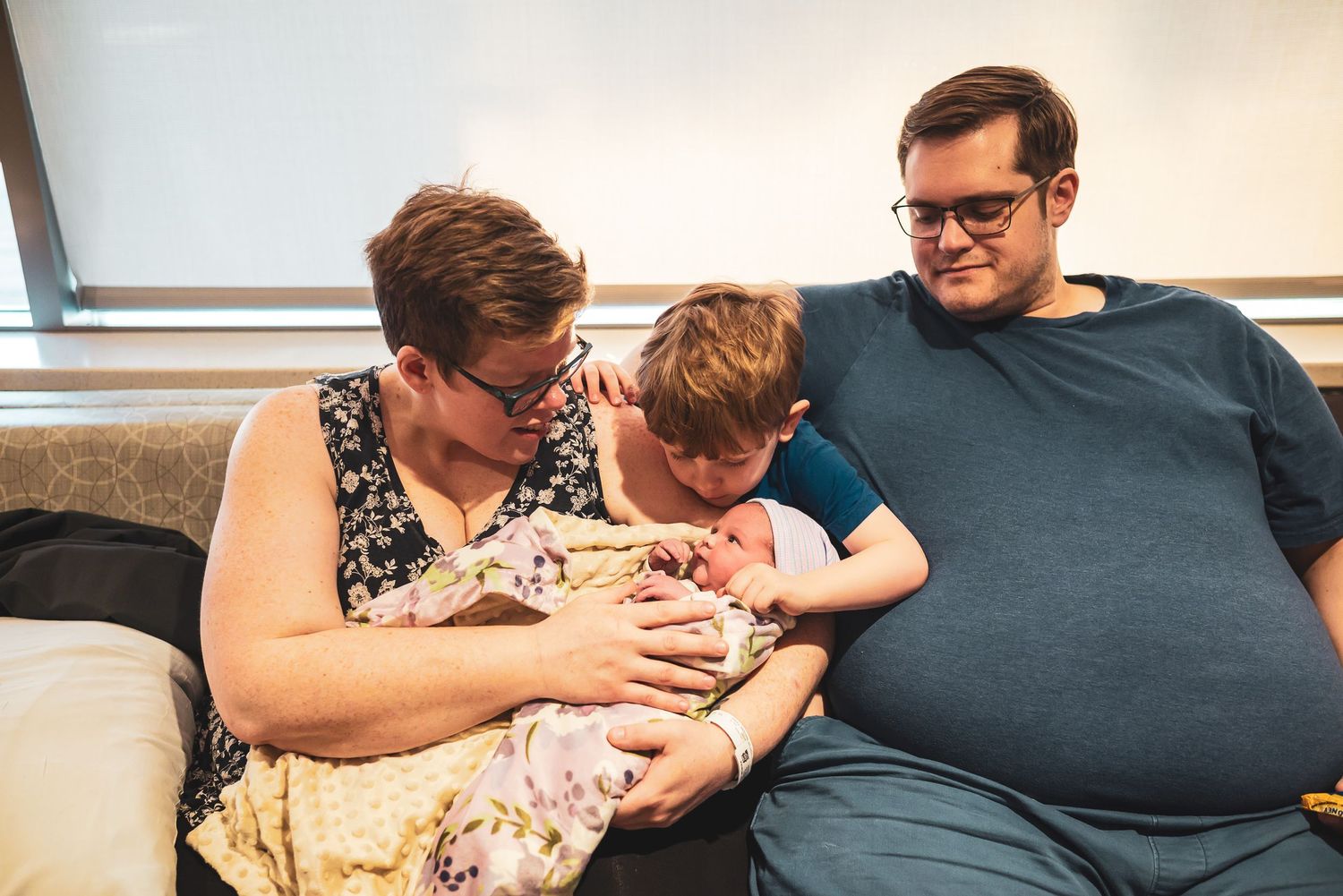 Three people sitting on a couch together look lovingly at a baby swaddled in blankets.