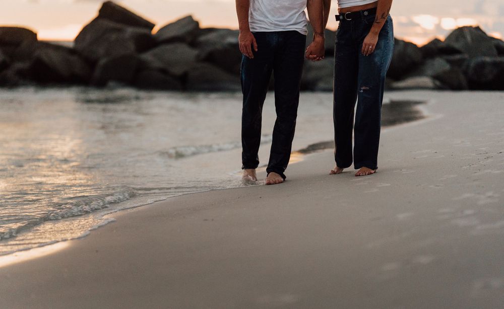 A couple walks hand in hand along a rocky beach at sunset with warm golden light reflecting on the water for engagement photoshoot Tampa FL