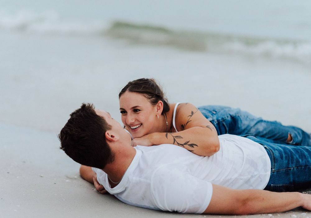 A couple wearing jeans laying down on the sand for engagement photoshoot by the beach with in St. Pete Florida
