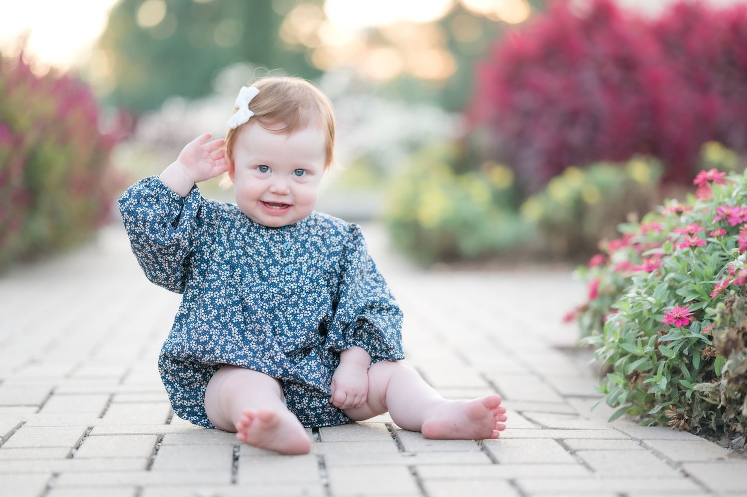 A one year old child in a white summer dress and wide-brimmed hat poses playfully in a garden with blooming flowers.