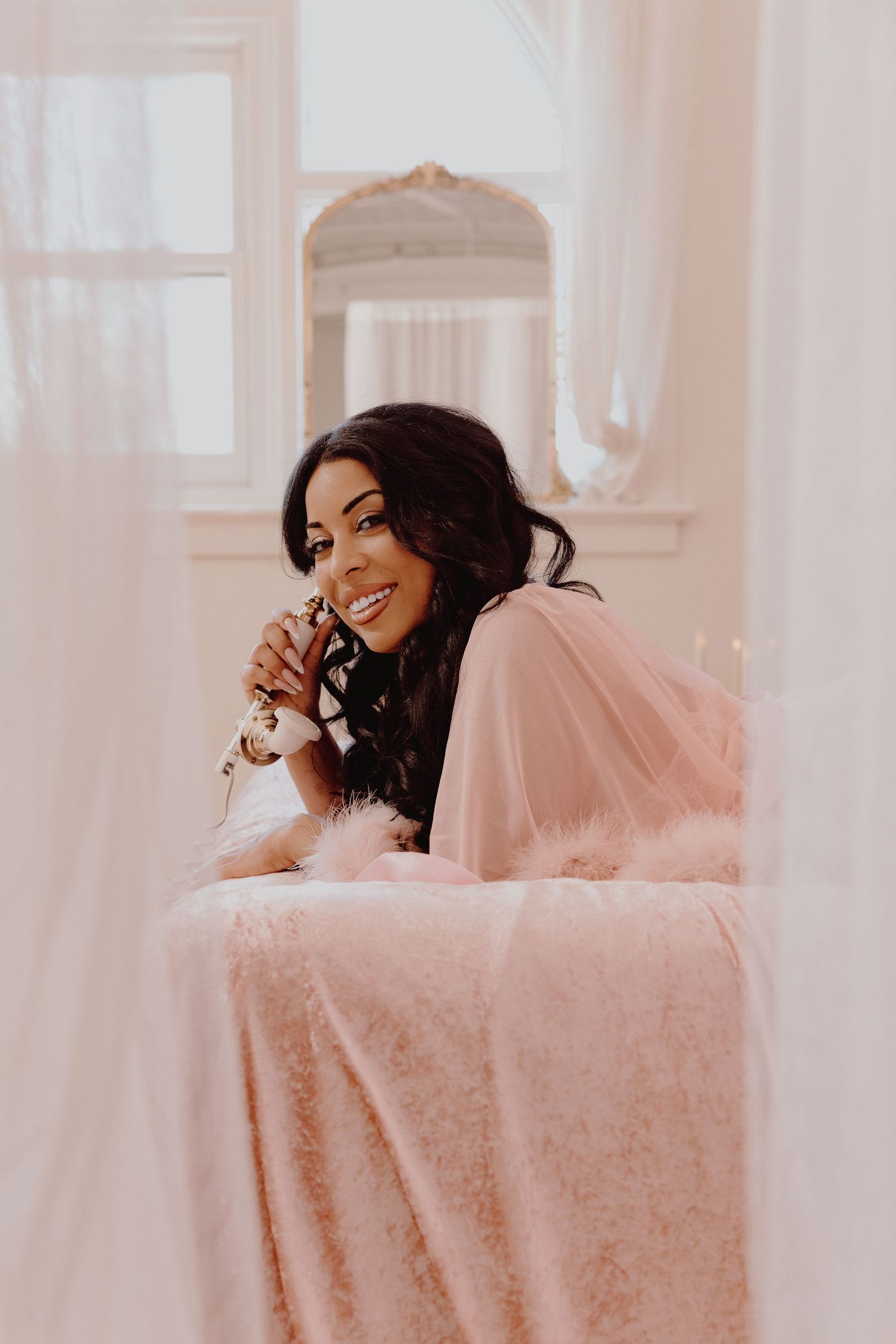 Smiling woman in pink tulle robe lying on pink fur bed holding champagne glass near bright window.