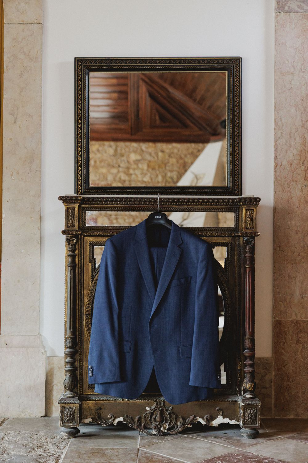 A navy blue suit groom jacket hangs on an ornate vintage console table with a decorative mirror above it.
