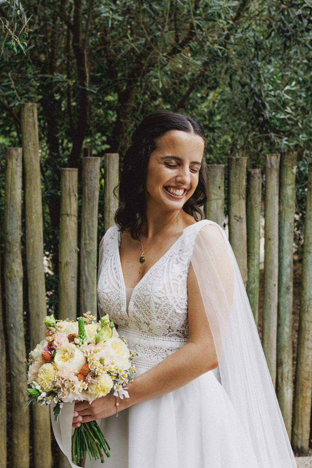 A bride in a lace V-neck wedding dress smiles while holding a soft pink and white floral bouquet in a garden setting in a wedding in lisbon