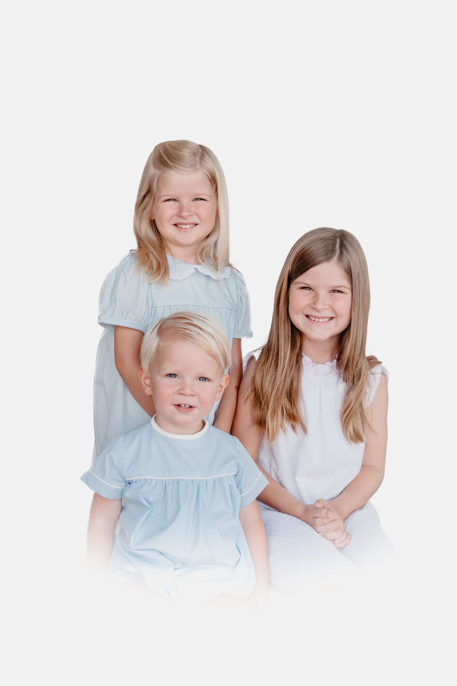 Three smiling blonde children in light blue and white dresses pose together for a portrait photo on white background.