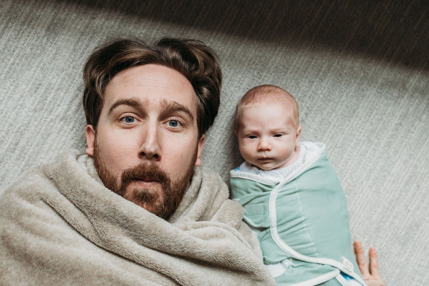 A bearded adult and swaddled baby lying on gray fabric looking up at the camera.
