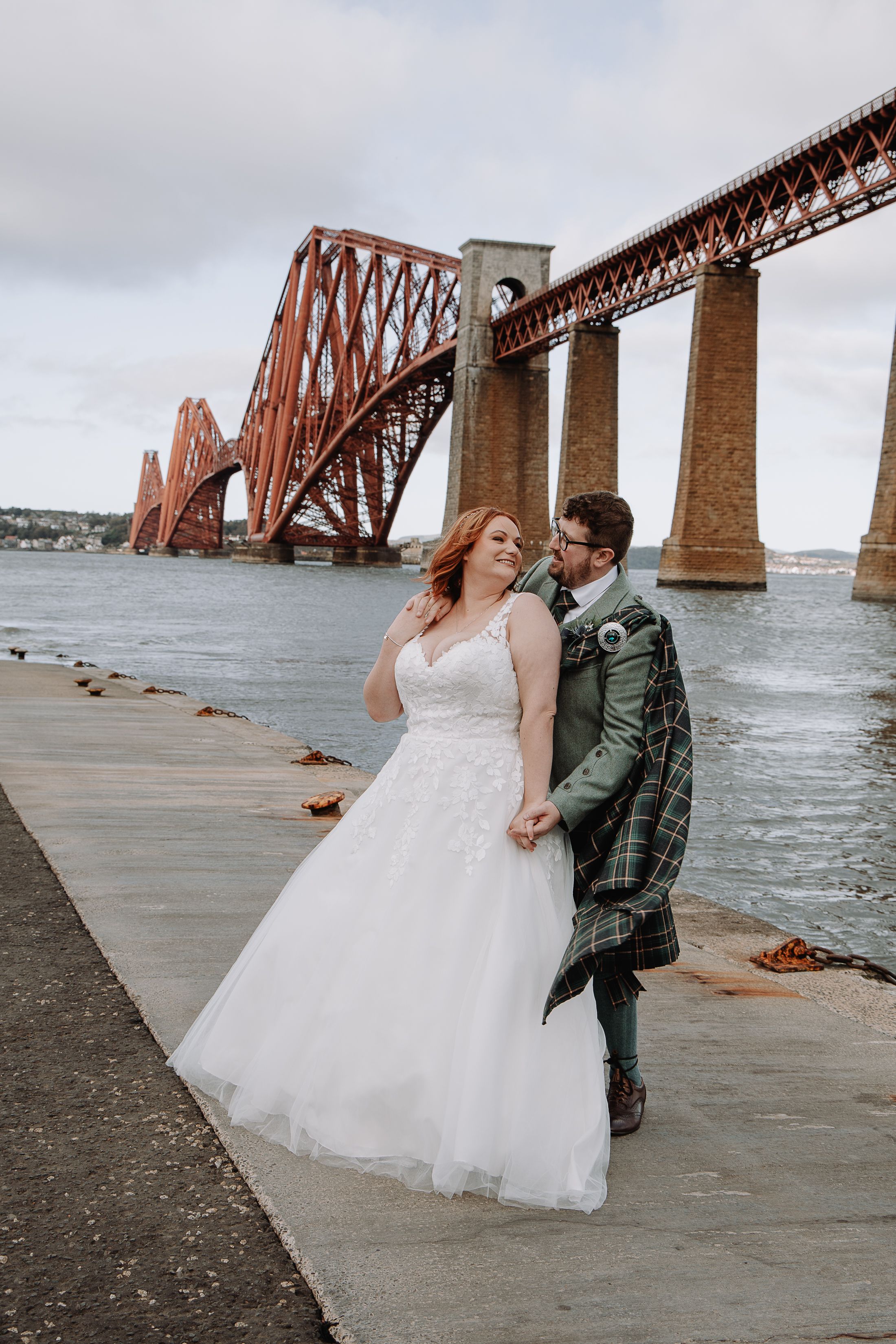 Newlywed couple posing by the historic Forth Bridge in Scotland.