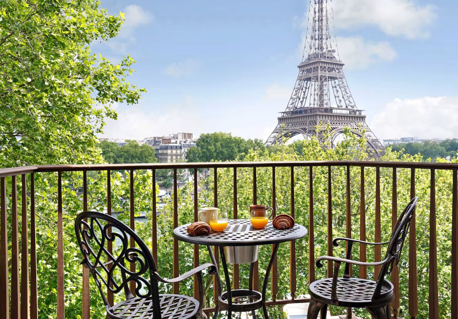 A romantic balcony view of the Eiffel Tower in Paris with a bistro table set for breakfast and green trees in summer.