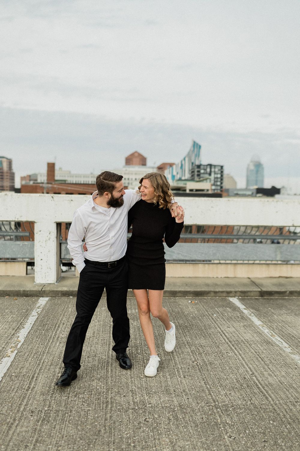 A couple embraces while standing on a rooftop parking deck with city buildings in the background.