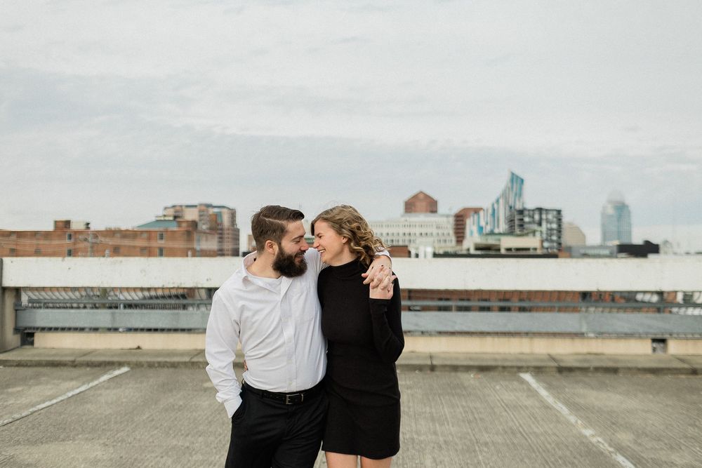 A couple stands together on an empty rooftop parking lot with urban buildings visible in the background.