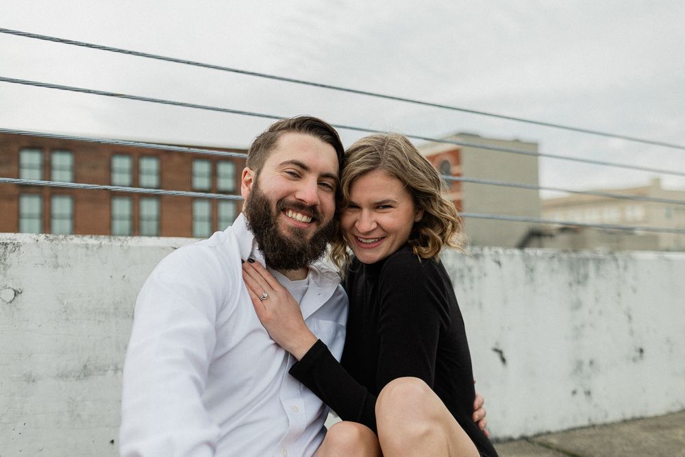 A couple laughs together during their rooftop photo session with urban architecture behind them.