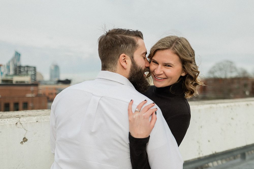 A couple embraces on an urban rooftop with city buildings visible in the background on an overcast day.