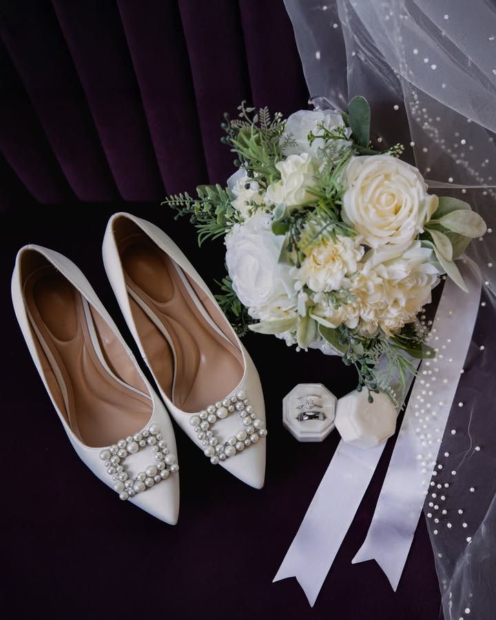 White bridal heels with pearl embellishments and floral bouquet arranged against a dark background.