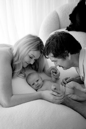 Black and white image of parents lying down and bonding with their newborn on white bedding.