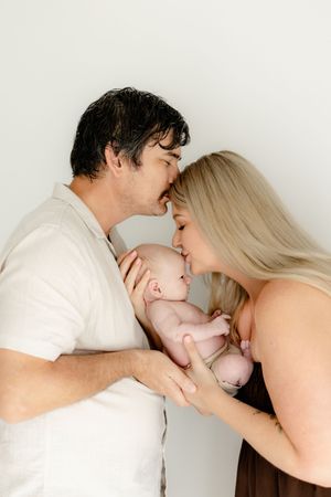Parents in white clothing sharing tender kisses with their newborn baby against a light background.