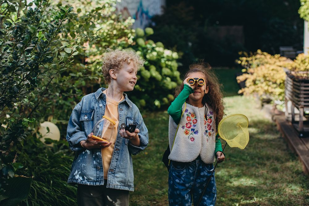 Two children play with bubble wands in a sunny garden, wearing casual denim and creating soap bubbles in the air.