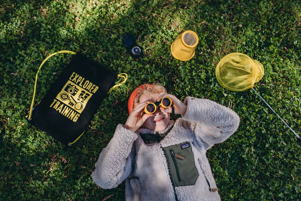 Person in a gray hoodie drinks from a yellow bottle against a green leafy background.