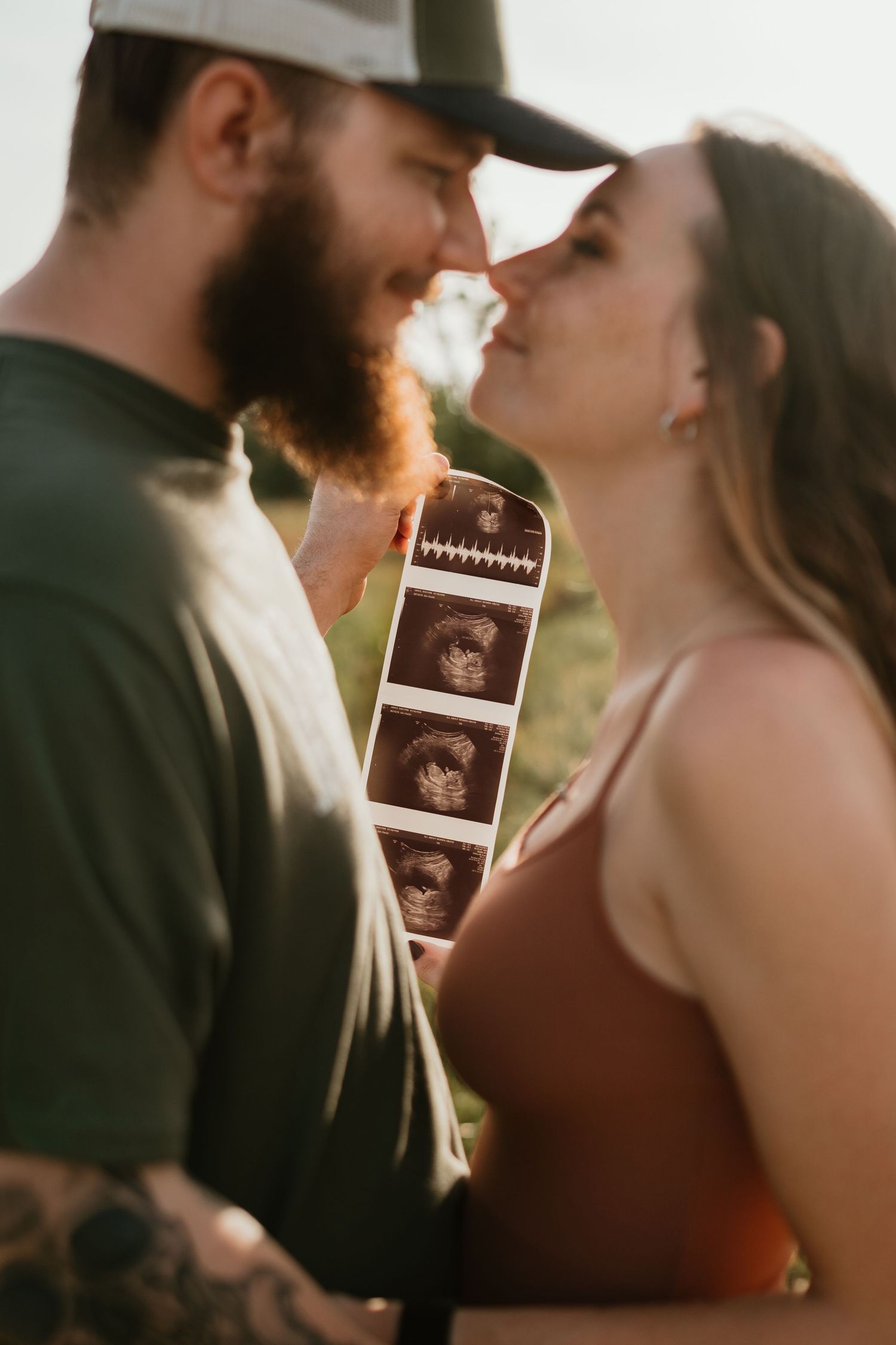 A couple shares an intimate moment while holding ultrasound photos in a golden sunset setting.