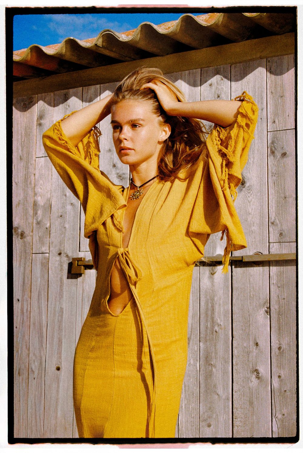Figure in a vibrant yellow dress poses against a rustic wooden wall in warm sunlight.