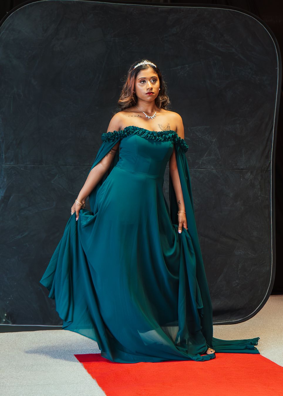 A model in a flowing teal evening gown poses dramatically against a dark backdrop in a photo series.