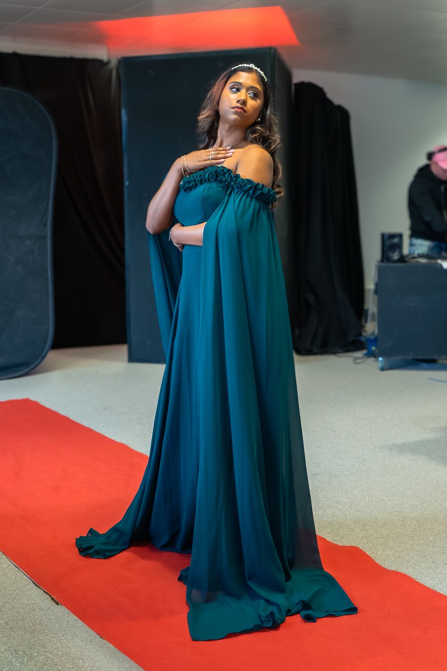 A model in a flowing teal evening gown poses dramatically on a red carpet during a professional photo shoot.