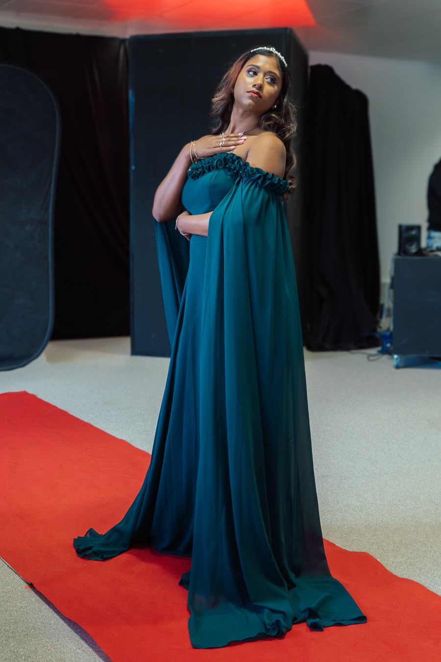 A dramatic fashion photoshoot featuring a flowing teal evening gown against a red carpet backdrop in a studio setting.