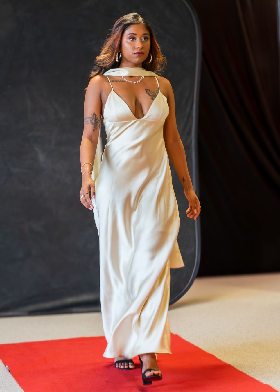 A person in a white slip dress walks confidently down a red carpet runway against a dark backdrop.