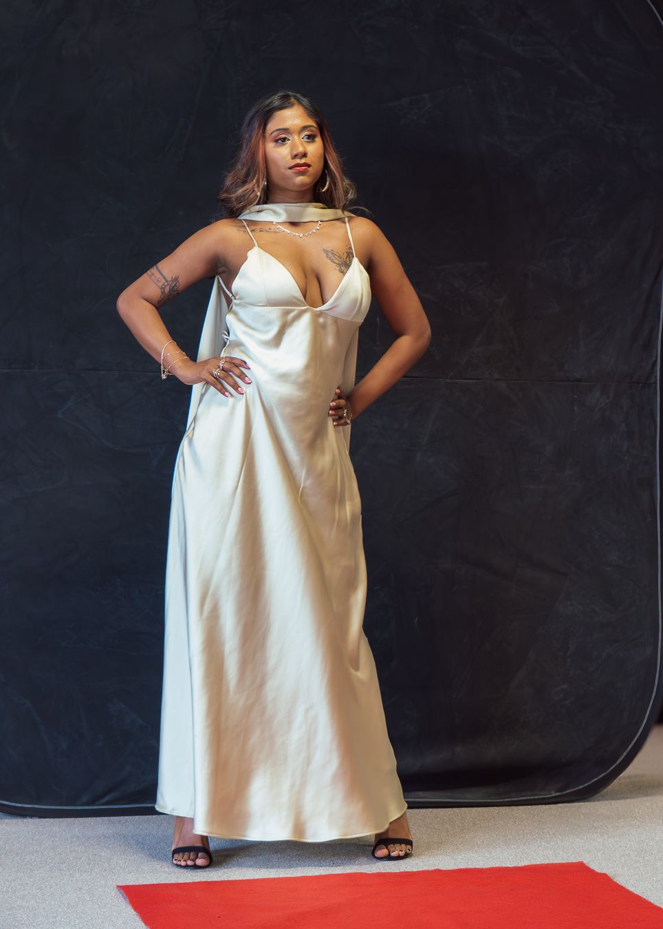 Model poses confidently in a flowing white satin evening gown with cutout details on a black backdrop with red carpet.