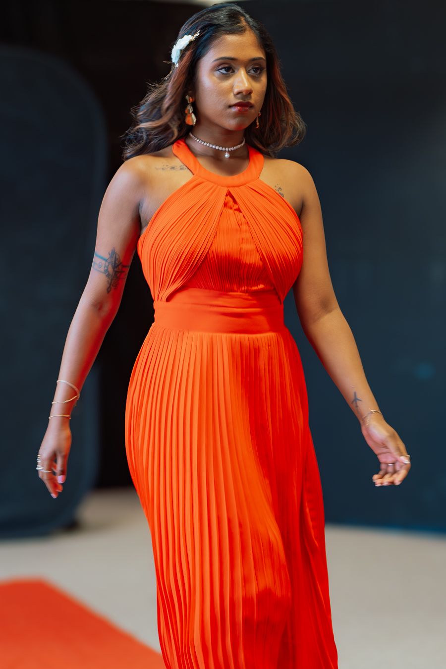 A model walks the runway in a vibrant orange pleated halter dress against a dark background.