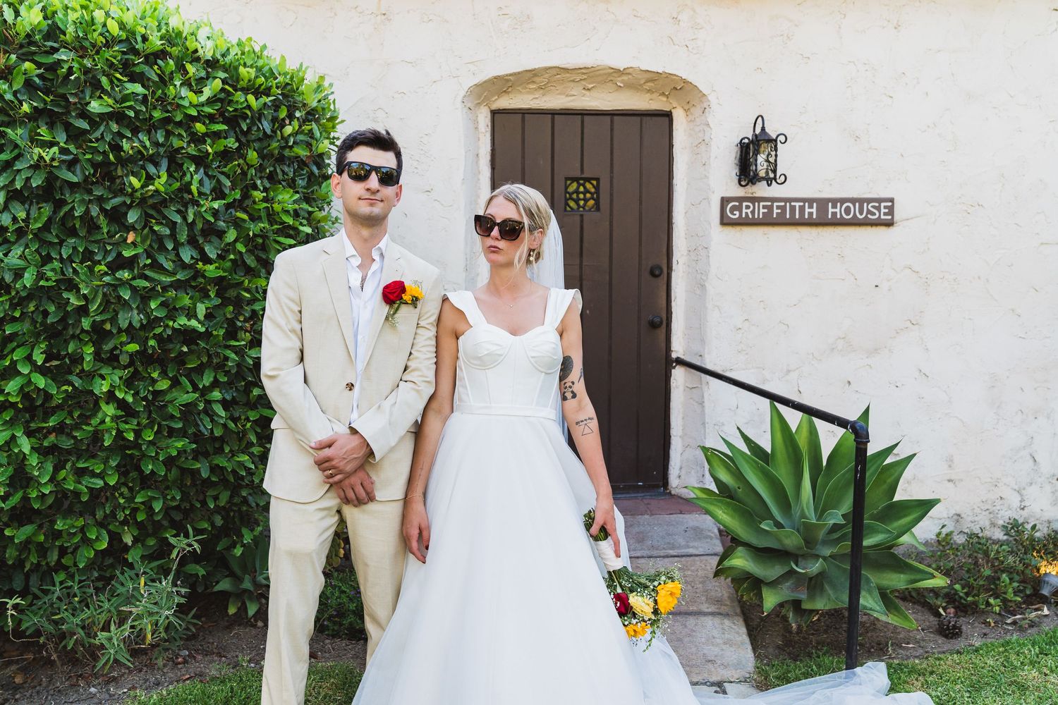 A wedding couple walks hand in hand through a garden path with lush greenery and white stucco walls.