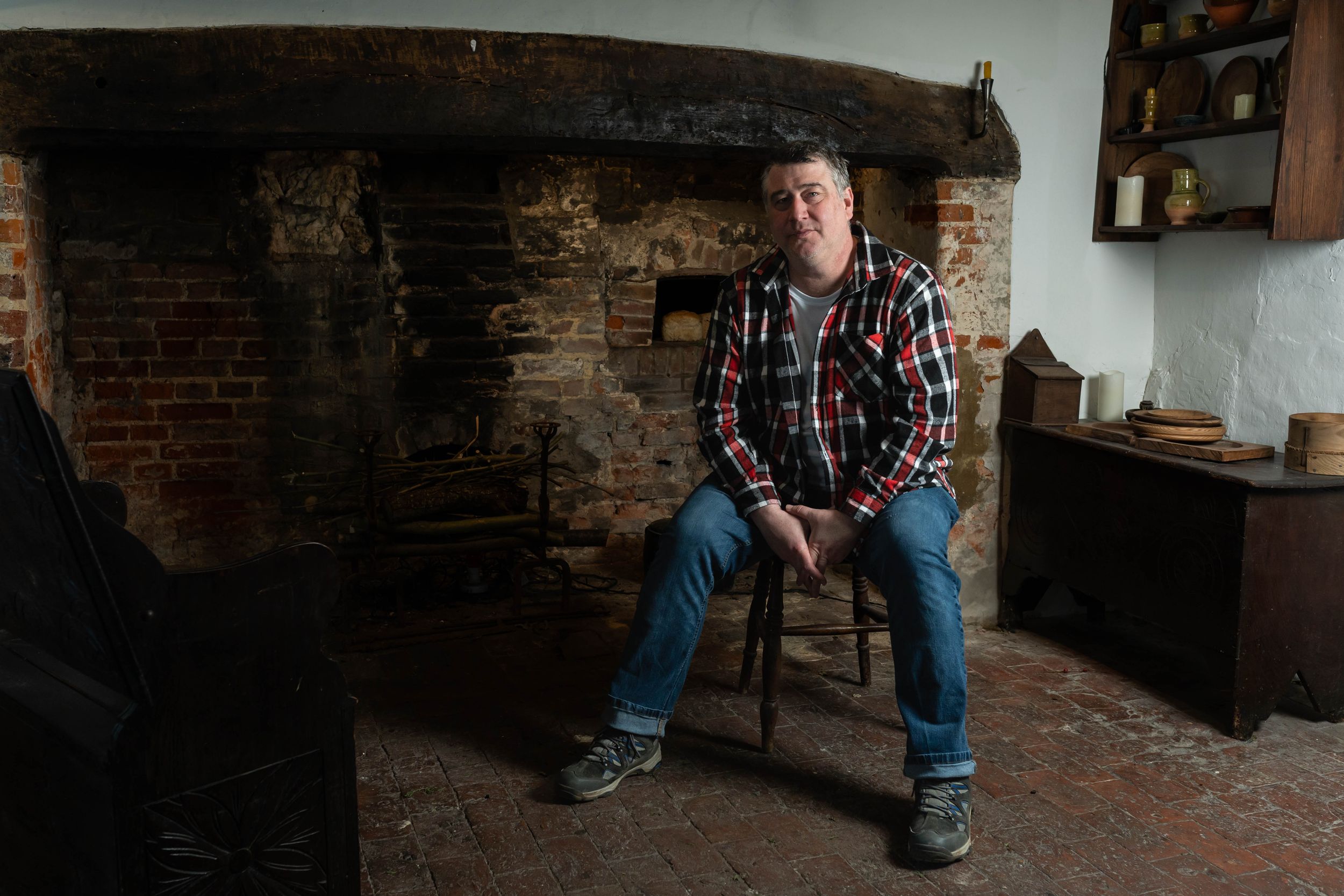Brighton theatre photography. an actor sits in a dimly lit cottage on a stool wearing jeans and a checked shirt