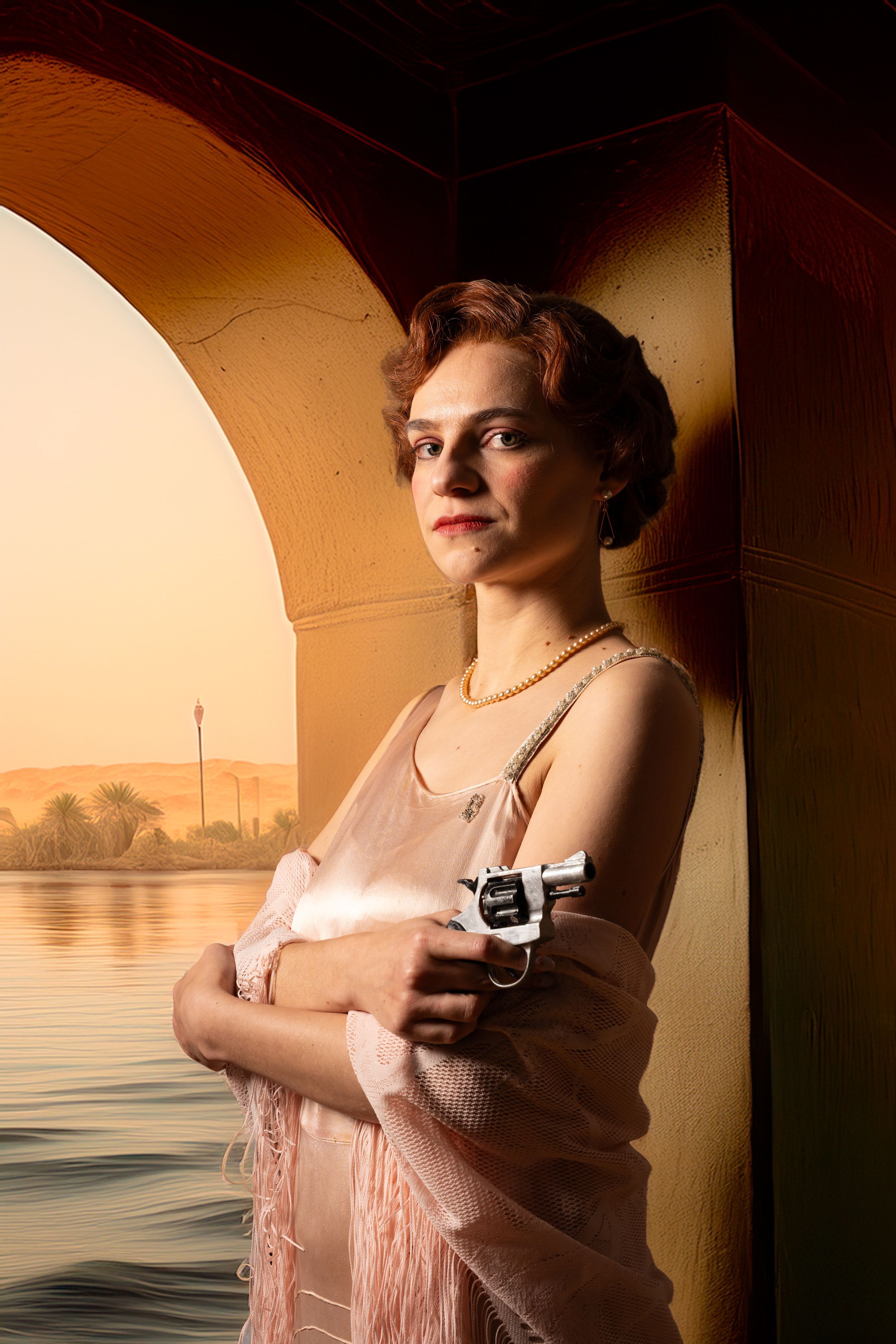 theatre photography promo shot Brighton of a woman in 1930s dress stands in an archway holding a gun with a sunset in the background