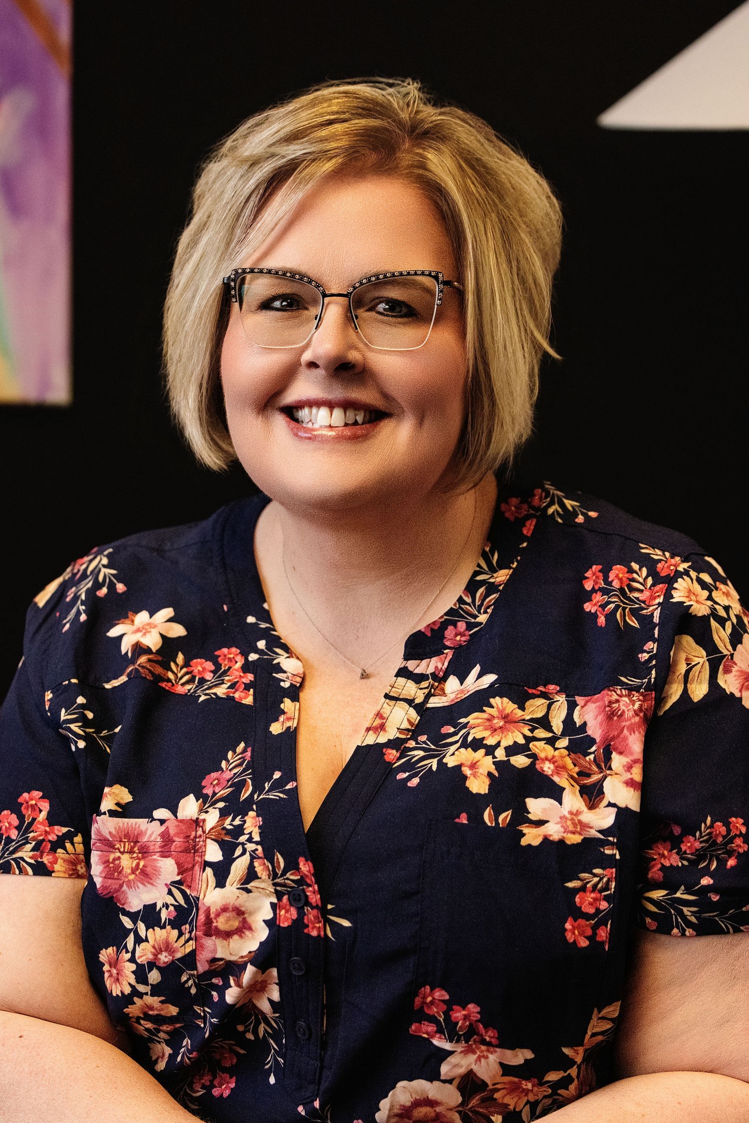 A person wearing glasses and a navy floral print dress smiles warmly in a professional portrait series.