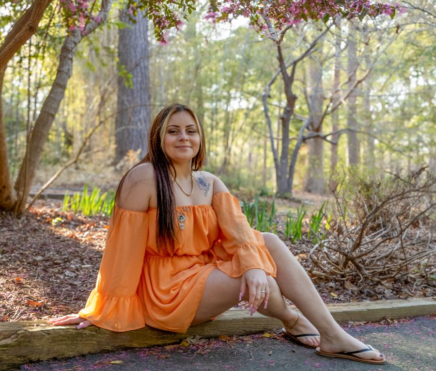 woman wearing an orange romper, sitting on the edge of a path with a wooden background with purple flowers from a tree above her