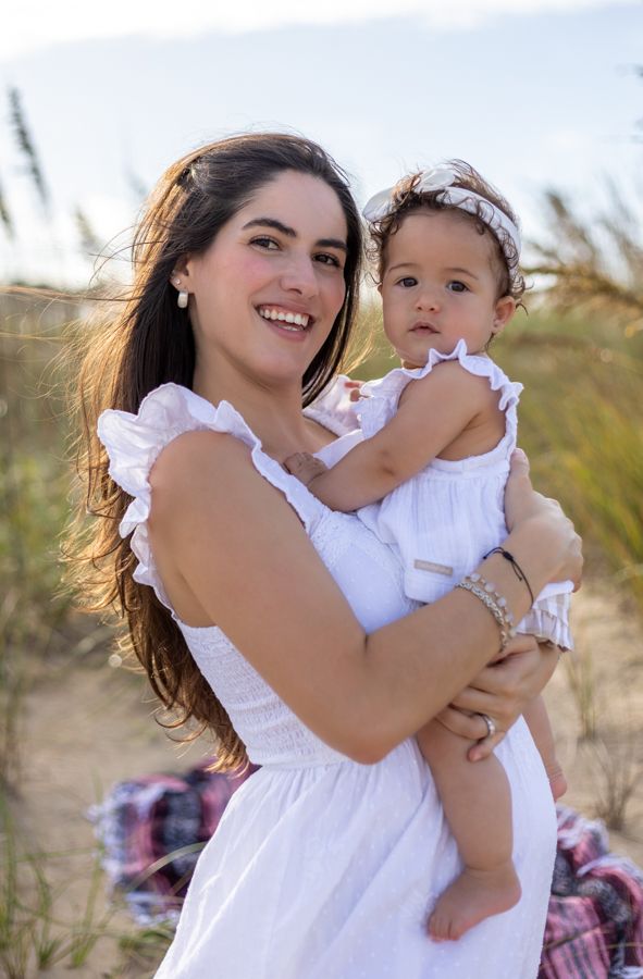 mom holding daughter on the beach, both wearing white with beach grass in th background