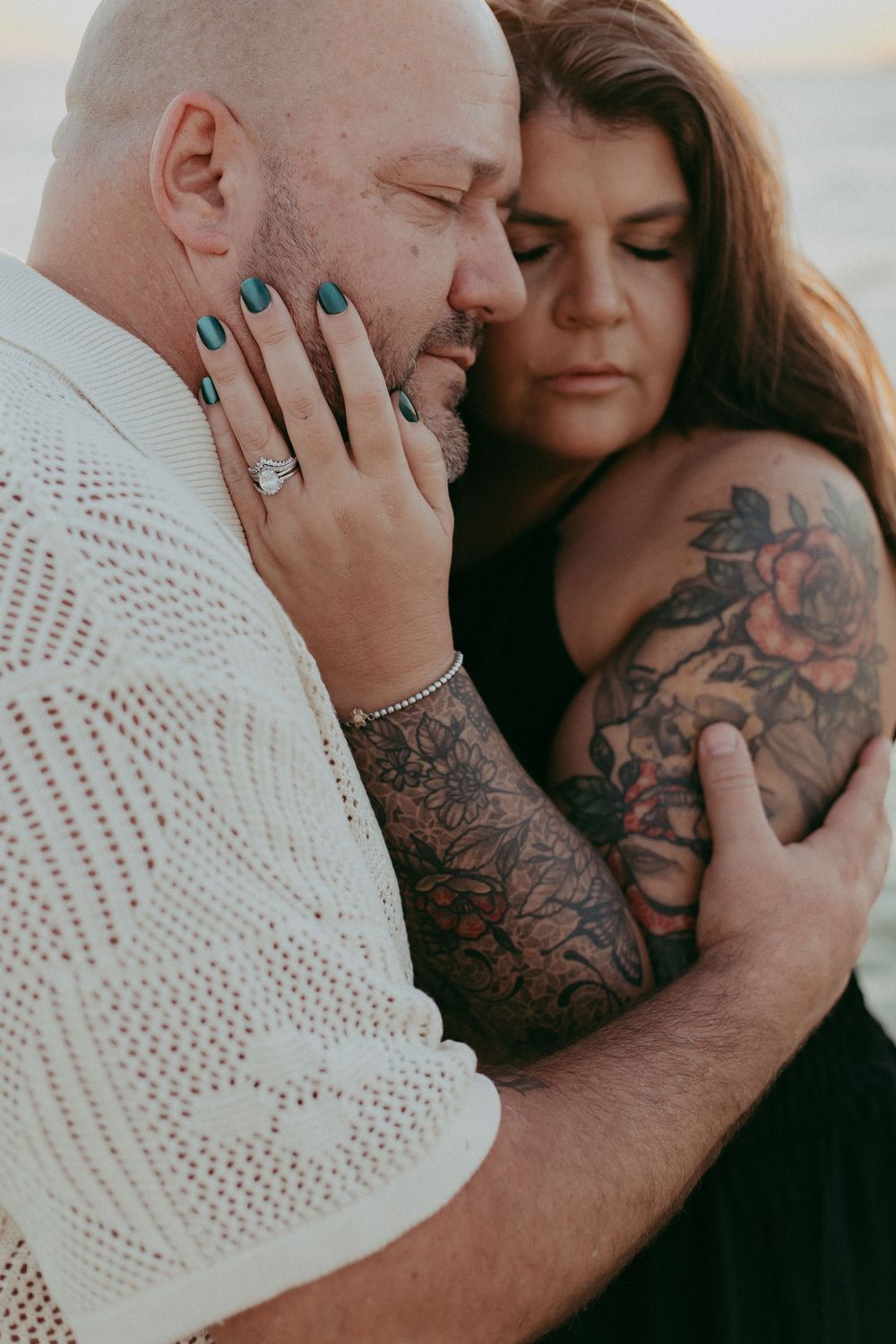 Intimate couple embraces in romantic portrait showing tattoos and engagement ring with teal nail polish.