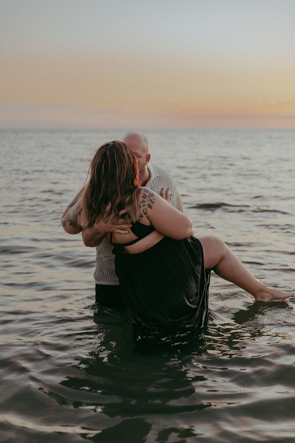 Two people embrace while wading in calm ocean waters at sunset creating a romantic silhouette against the horizon.